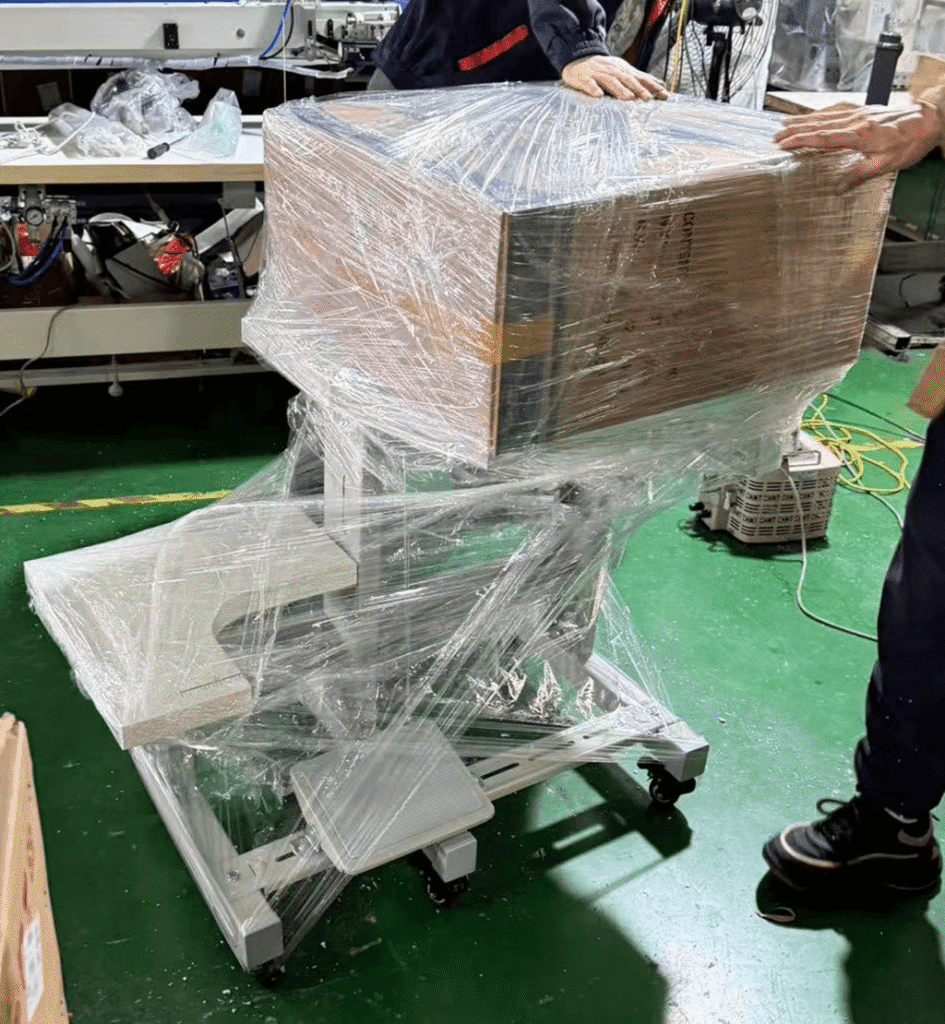 Industrial device on a wheeled stand wrapped in plastic wrap, hands securing the top in a factory setting.