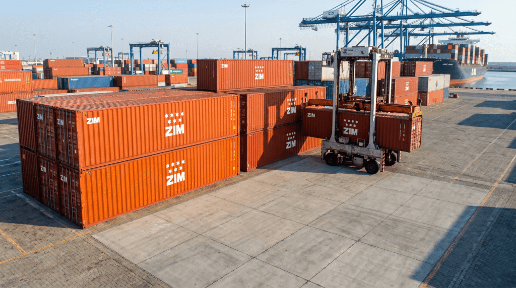Port scene with stacked orange ZIM shipping containers, a container handler, blue gantry cranes, and a cargo ship in the background.