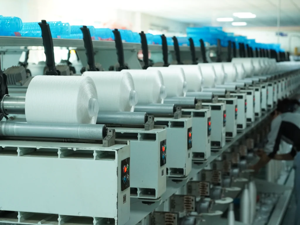Row of industrial textile machines with white yarn on rollers in a factory setting blue bins above